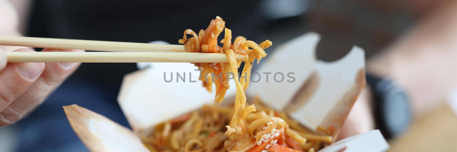 A man eats Chinese food from a box with wooden chopsticks, close-up. Traditional asian fast food, fresh delicious lunch