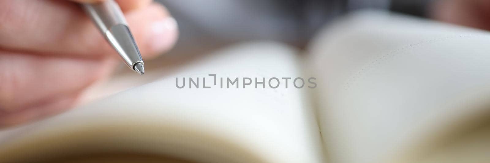 A woman's hand holds a pen near a notepad close-up, blurry. Document signature, synopsis, research and training