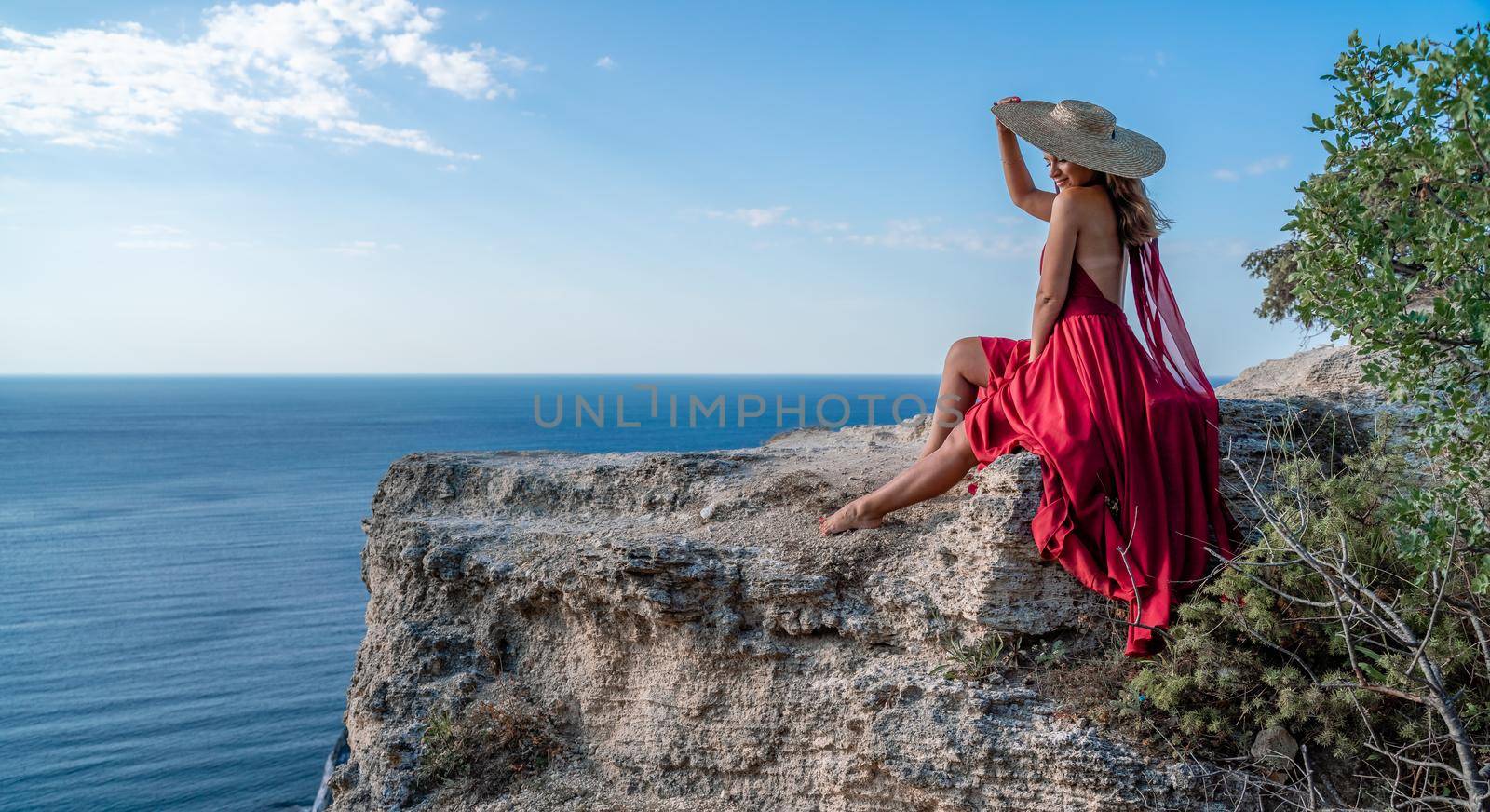 A girl with flowing hair in a long red dress sits on a rock above the sea. The stone can be seen in the sea. by Matiunina