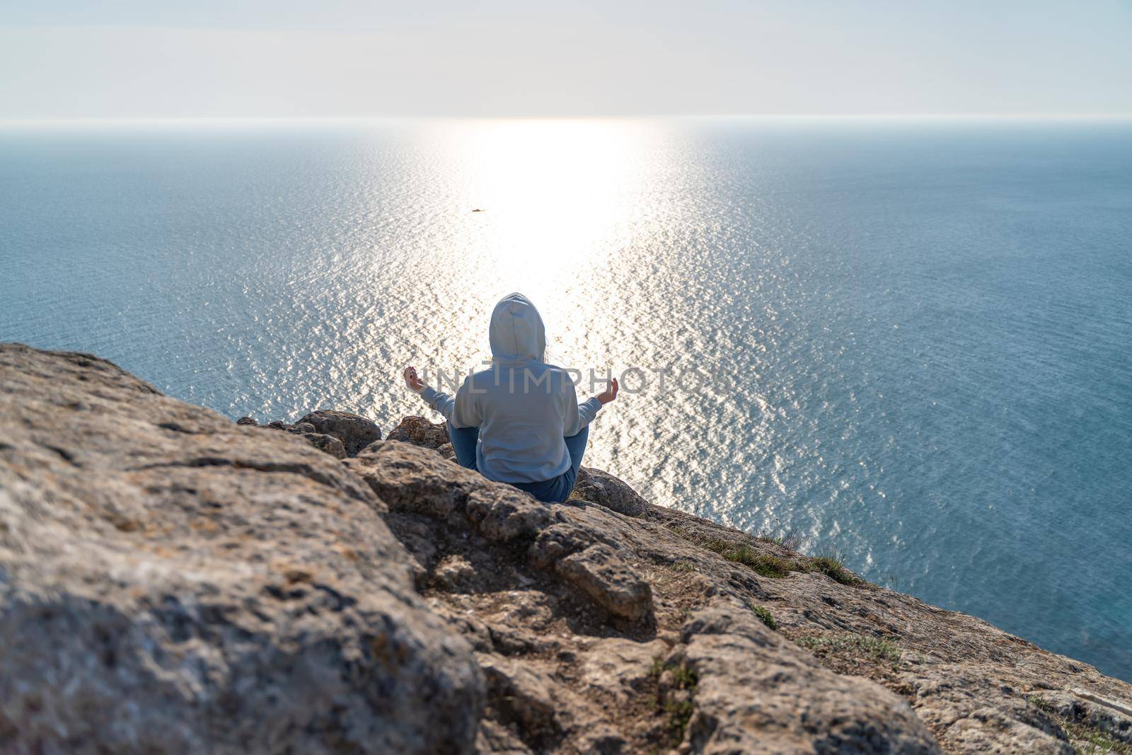 Woman tourist enjoying the sunset over the sea mountain landscape. Sits outdoors on a rock above the sea. She is wearing jeans and a blue hoodie. Healthy lifestyle, harmony and meditation.