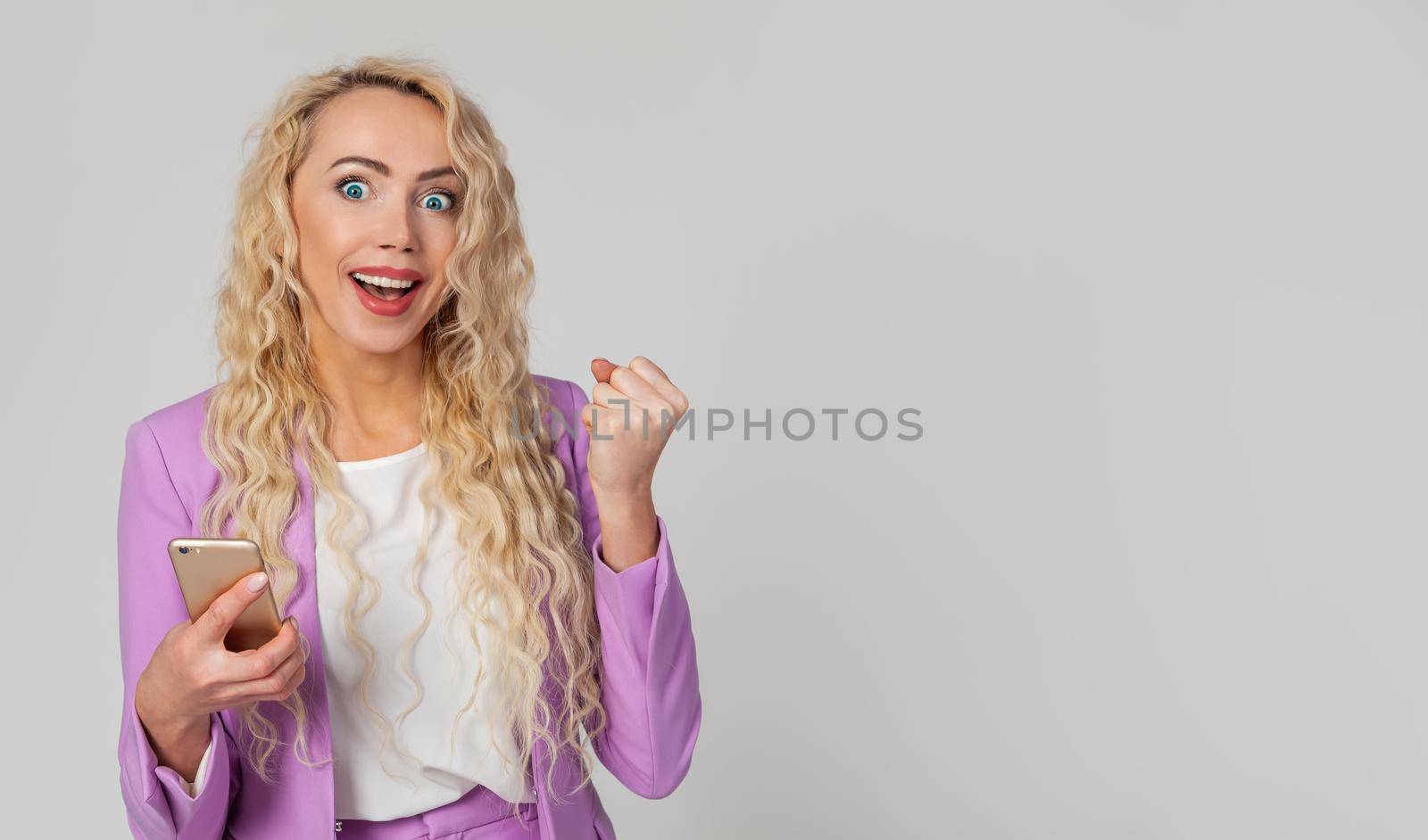 Happy triumphant woman holds a smartphone, fist and an enthusiastic smile, receives great fantastic news using a text message on her mobile phone. Wins the online lottery, receives the first prize.