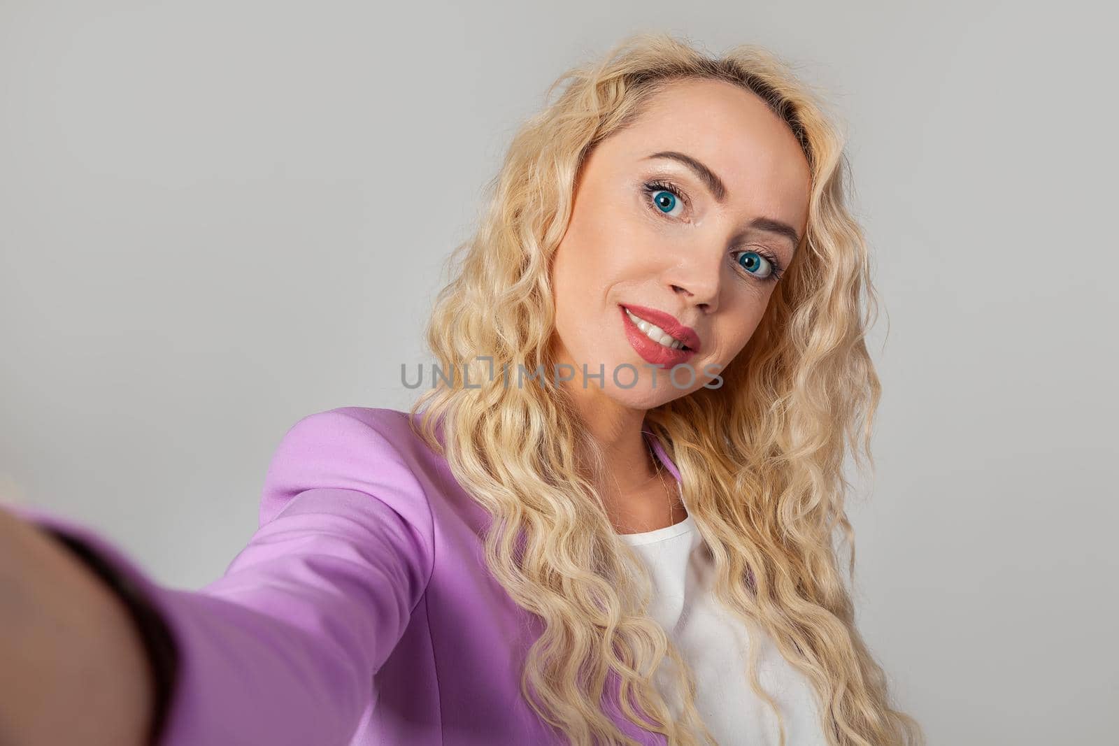 Closeup portrait of a stylish beautiful blonde woman with blue eyes, taking a selfie on a smartphone, video calling friends through an application, pleased with a smile, white background
