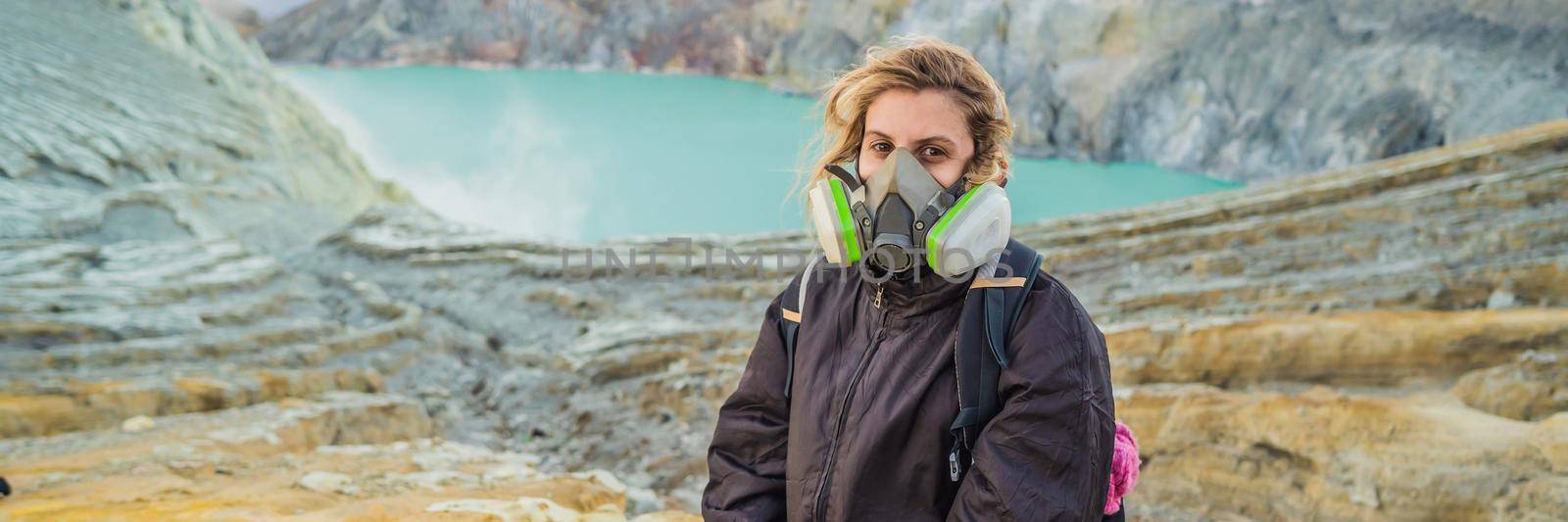 BANNER, LONG FORMAT Young woman tourist sitting at the edge of the crater of the Ijen volcano or Kawah Ijen on the Indonesian language. Famous volcano containing the biggest in the world acid lake and sulfur mining spot at the place where volcanic gasses come from the volcano. She wears a respirator to protect from dangerous volcanic gasses by galitskaya
