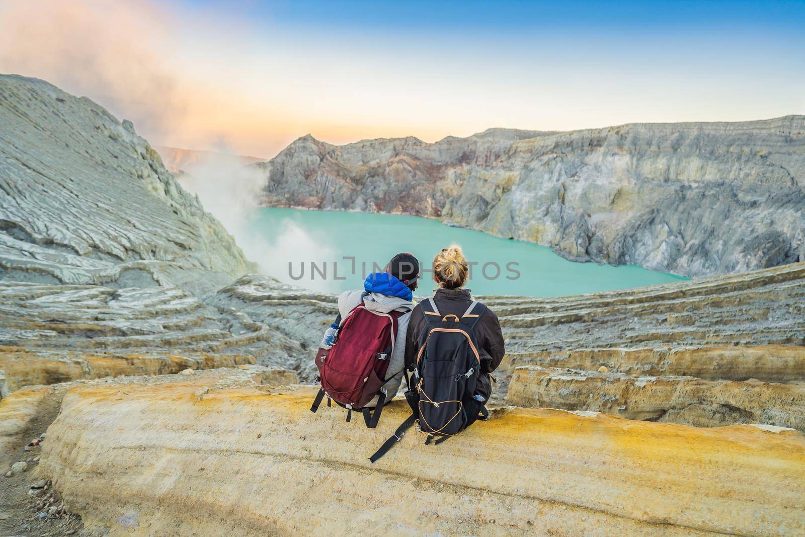 Young tourist man and woman sit at the edge of the crater of the Ijen volcano or Kawah Ijen on the Indonesian language. Famous volcano containing the biggest in the world acid lake and sulfur mining spot at the place where volcanic gasses come from the volcano. They enjoy the magnificent view on the crater.