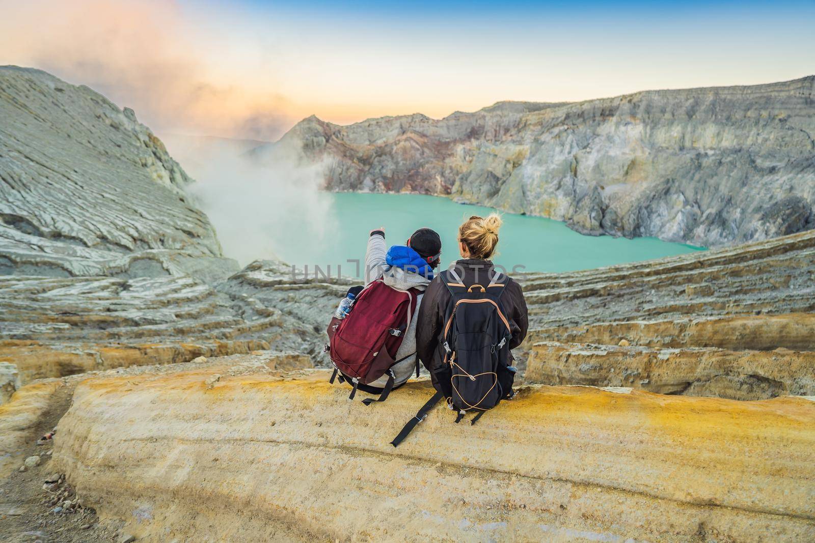 Young tourist man and woman sit at the edge of the crater of the Ijen volcano or Kawah Ijen on the Indonesian language. Famous volcano containing the biggest in the world acid lake and sulfur mining spot at the place where volcanic gasses come from the volcano. They enjoy the magnificent view on the crater.