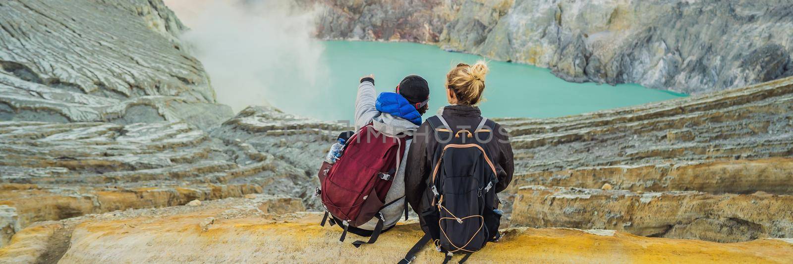 BANNER, LONG FORMAT Young tourist man and woman sit at the edge of the crater of the Ijen volcano or Kawah Ijen on the Indonesian language. Famous volcano containing the biggest in the world acid lake and sulfur mining spot at the place where volcanic gasses come from the volcano. They enjoy the magnificent view on the crater.