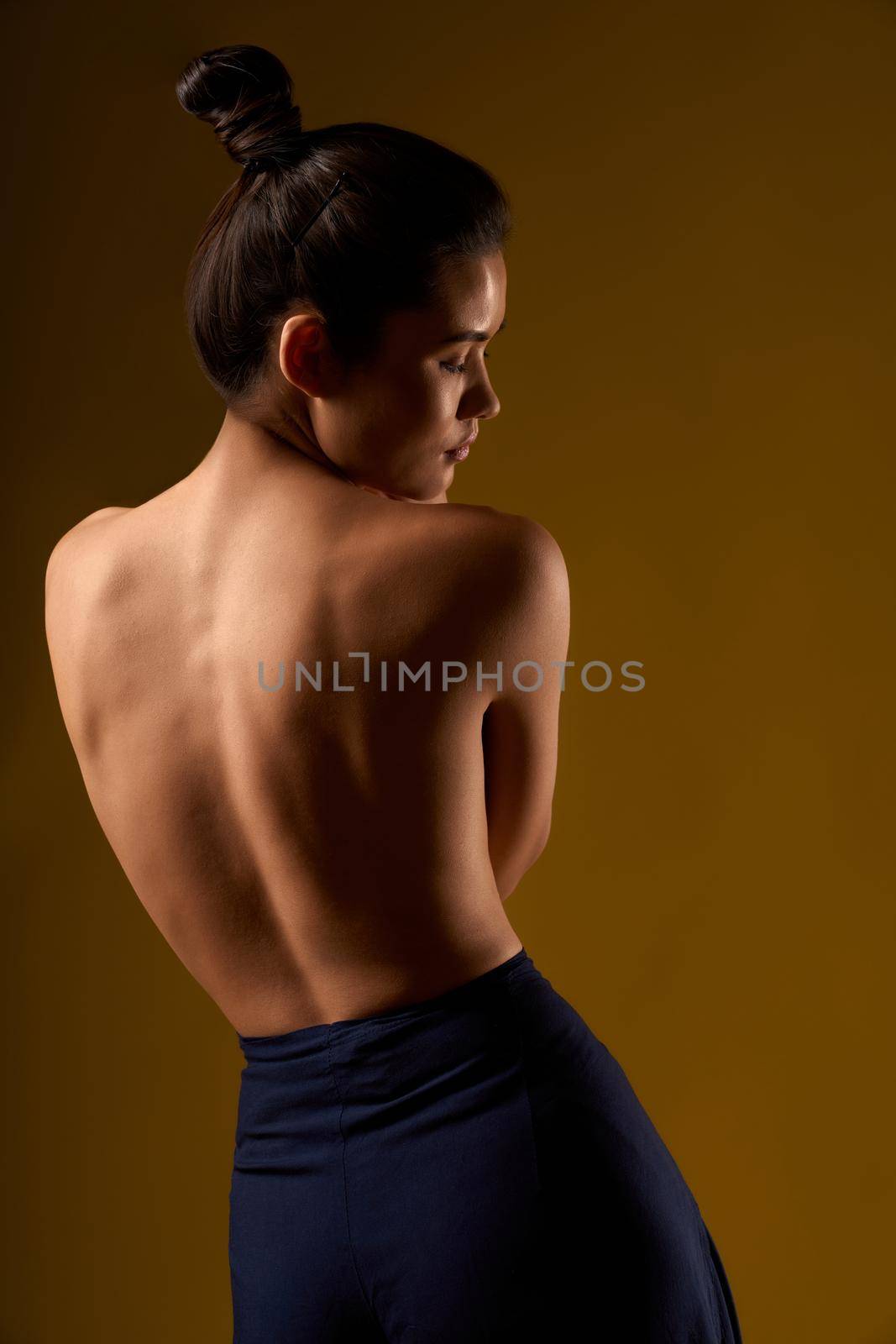 Back view of slim girl with hair bun standing, bending, looking down. Flexible female with bare back holding by hands, breezing, meditating, Concept of yoga doing and new age.