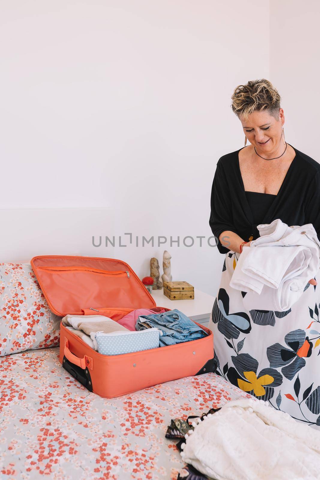 mature woman folding clothes to prepare her luggage. woman owner of establishment preparing her suitcase for business trip. copy space. concept of holidays, travel. bedroom with natural light, uncluttered environment. with white walls.