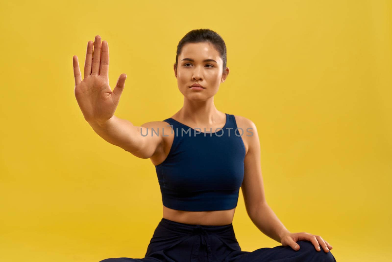 Front view of young female sitting, holding hand, looking forward. Sporty brunette wearing blue top, meditating, practicing yoga pose. Isolated on yellow studio background.