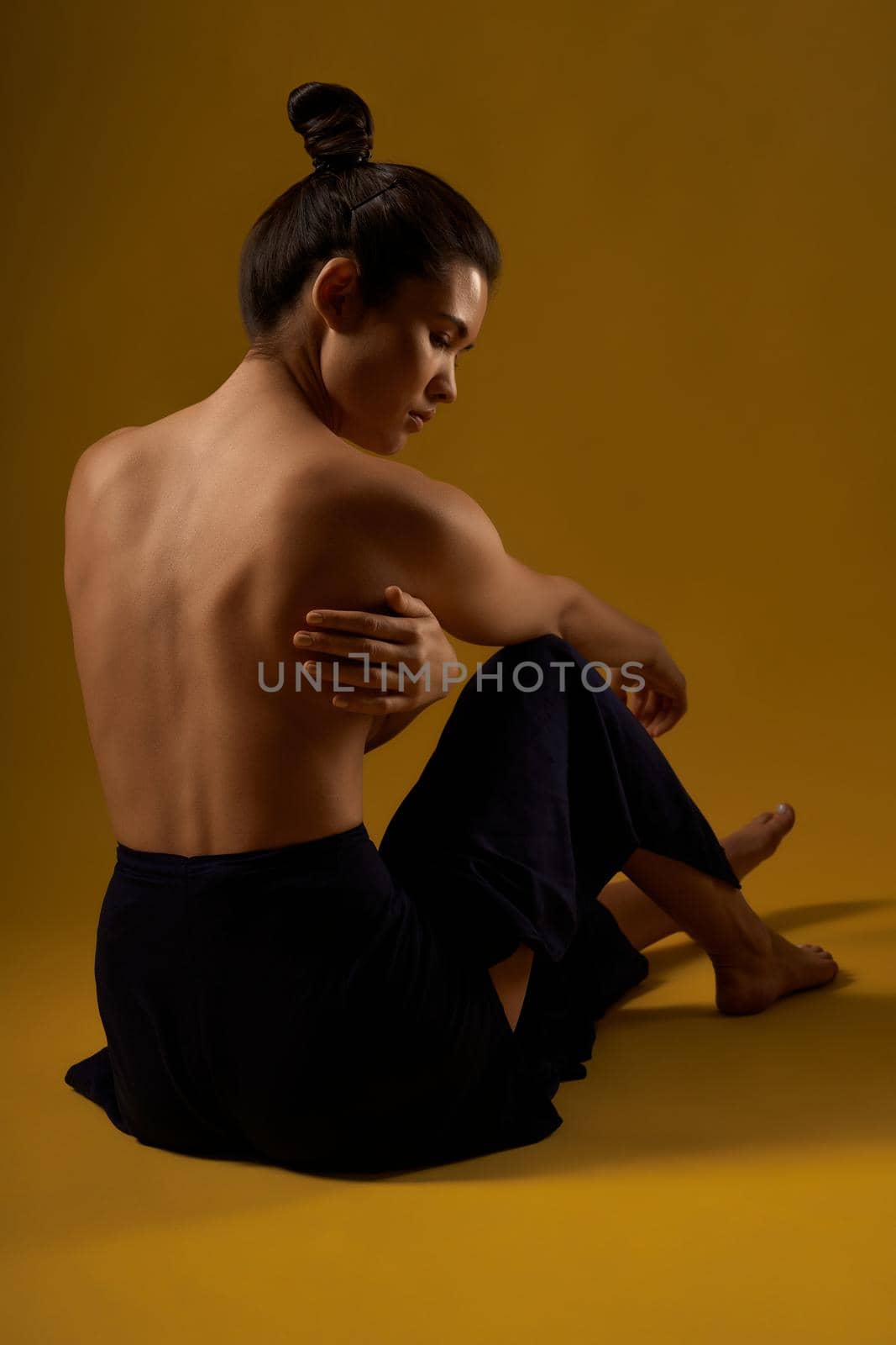 Back view of young sporty woman sitting backwards on floor, touching bare back by hand. Pretty brunette with hair bun practicing yoga. Concept of new age and healthy lifestyle.