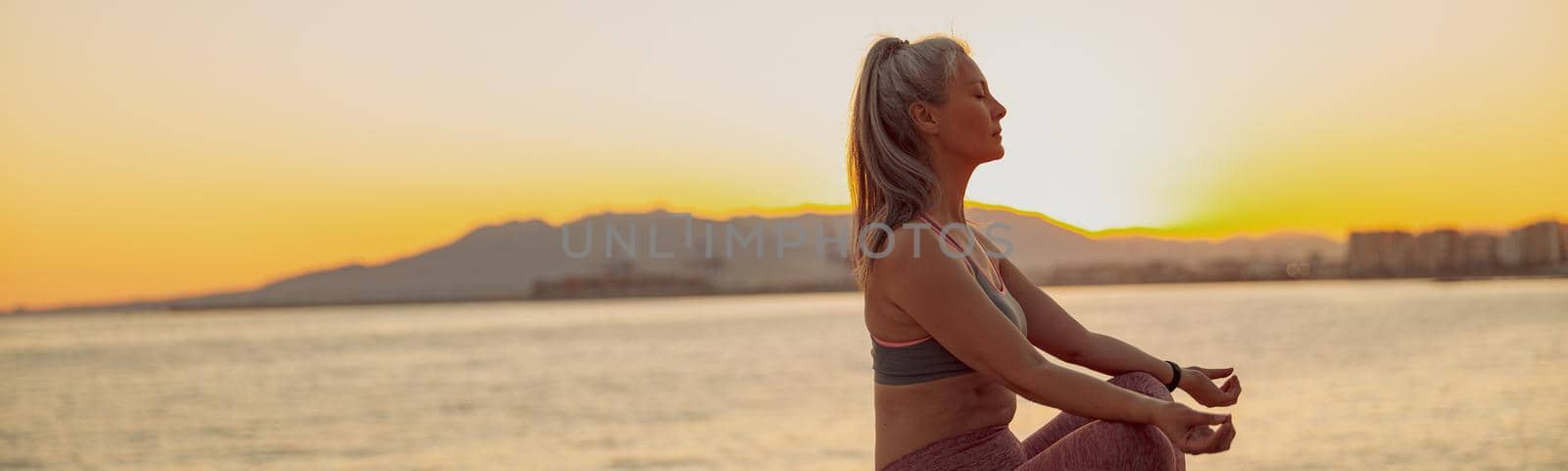 Side view of sporty lady practicing yoga, sitting on edge at seaside on background of sky at sunset and mountains