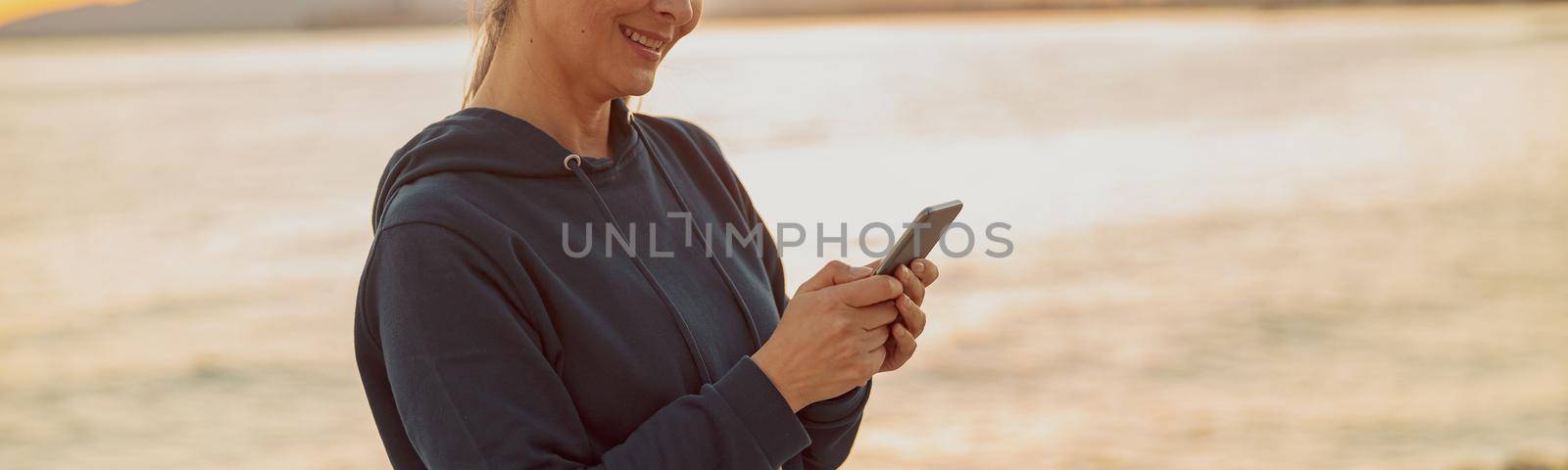 Charming sportswoman using smartphone and listening to music while standing on background of calm sea and sunset