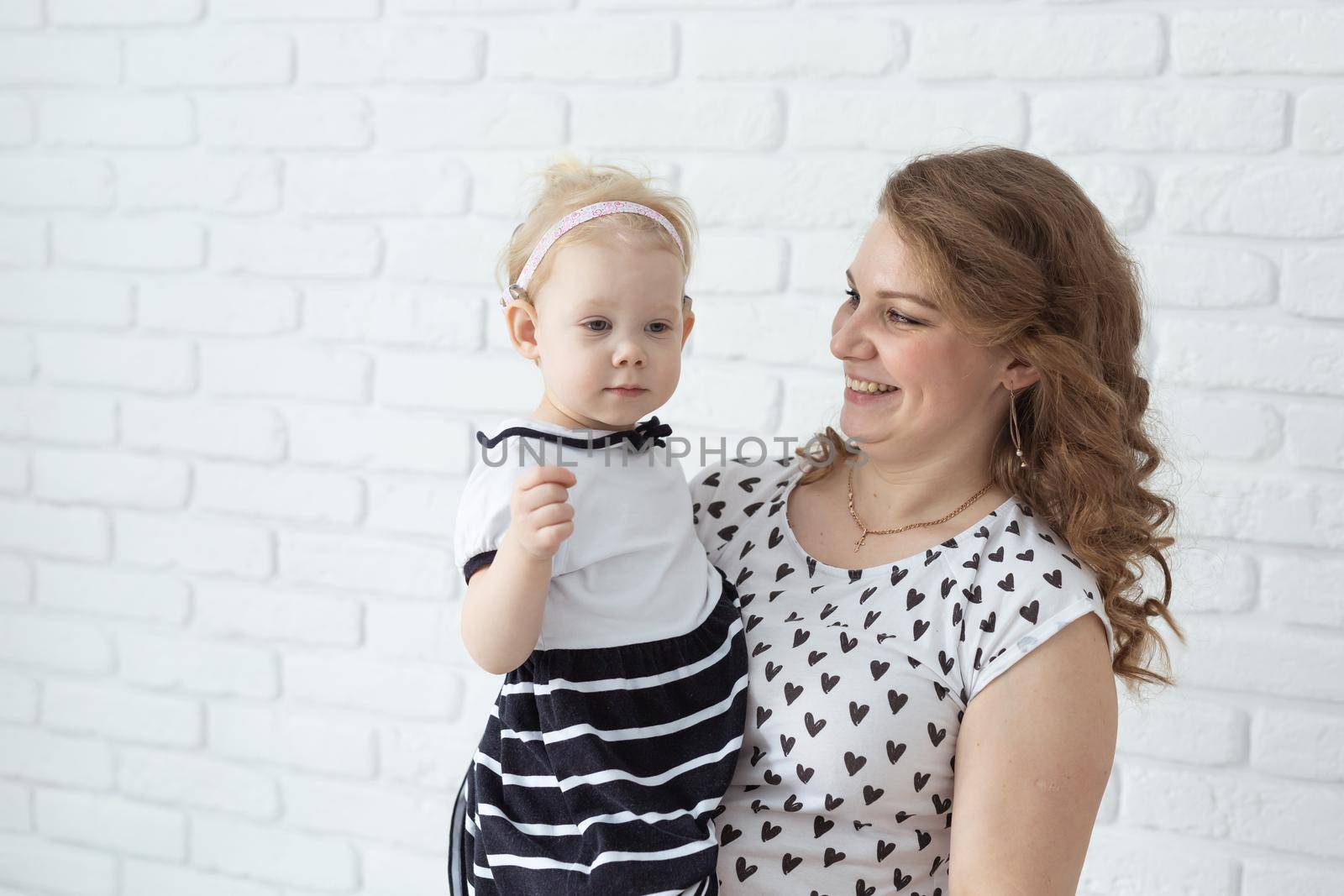 Mother holds and having fun her child with hearing aids and cochlear implants on white brick wall with copy space and place for advertising . Deaf and innovating medicine technologies concept by Satura86