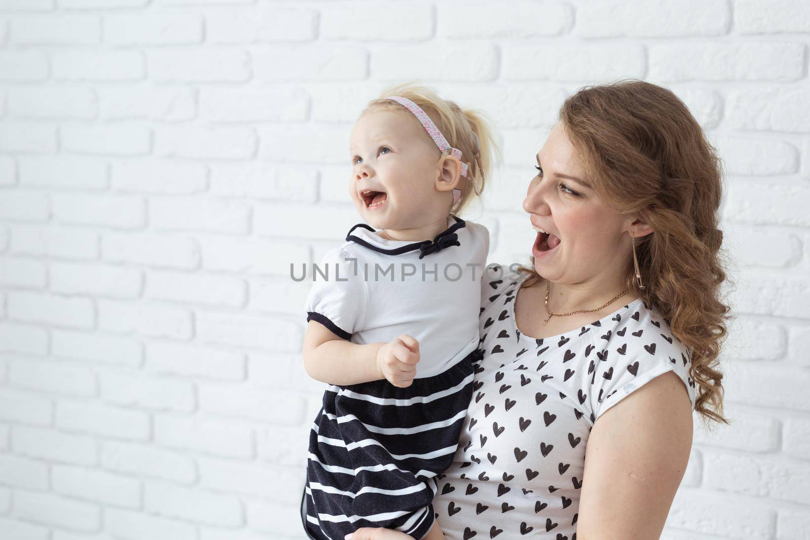 Mother holds and having fun her child with hearing aids and cochlear implants on white brick wall with copy space and place for advertising . Deaf and innovating medicine technologies concept by Satura86