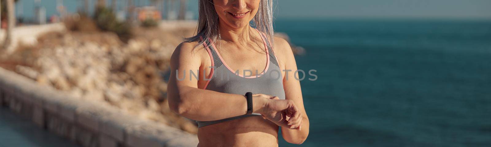 Active adult lady in sports top and leggings standing by the seashore and looking at fitness bracelet