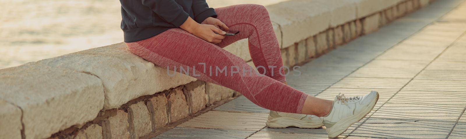 Photo of woman legs in pink leggings and white sneakers sitting on seashore by the sea and holding smartphone in hands