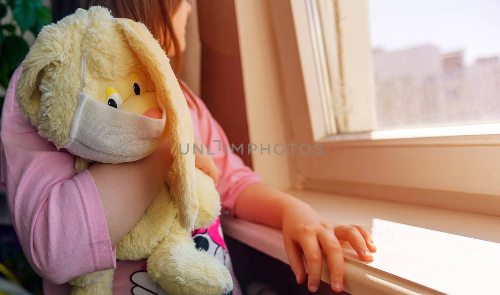 A girl with her toy, a plush rabbit, in a mask looks out the window concept of protection against coronavirus, quarantine, stay at home.