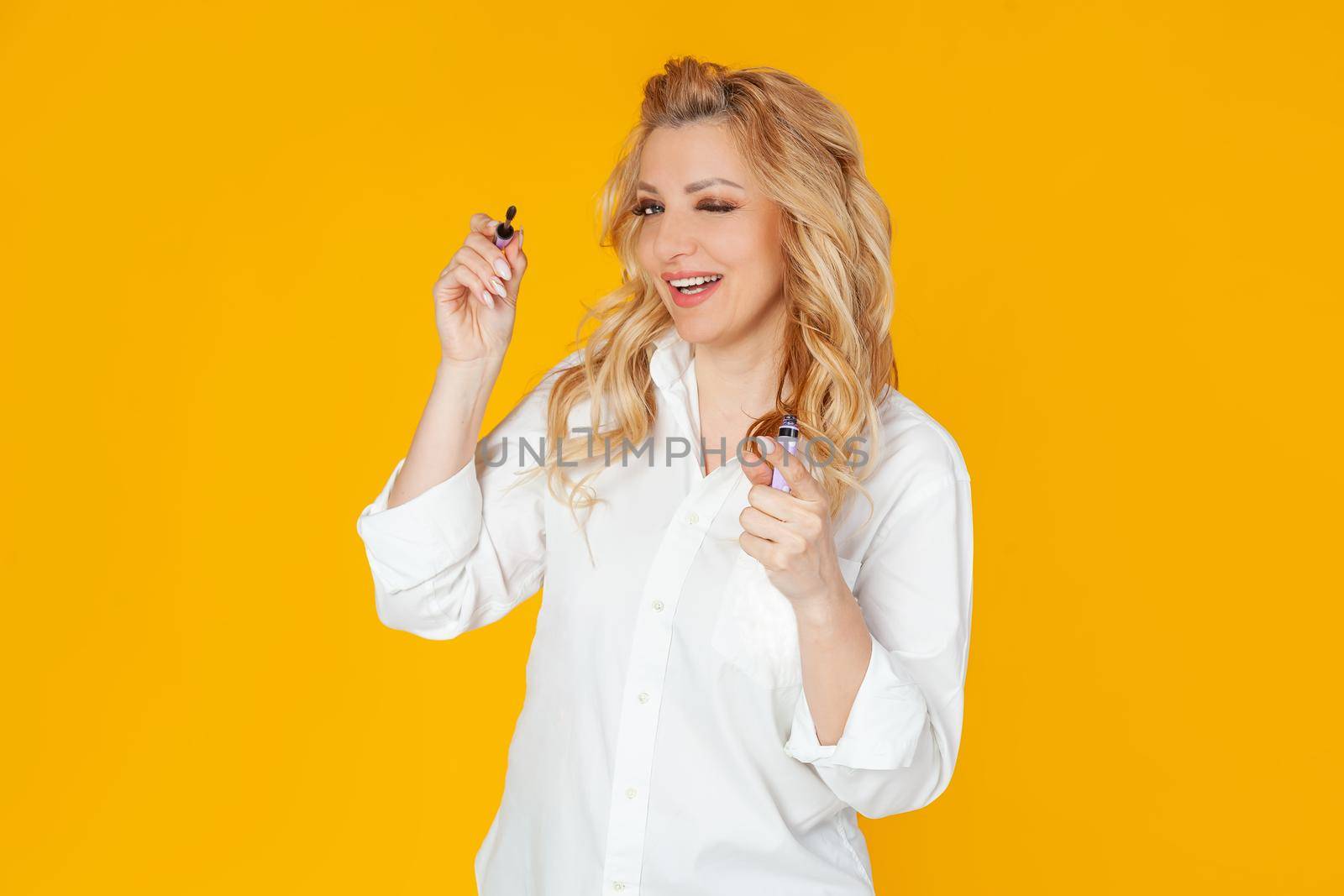 Blond in white shirt, middle aged European woman winks and smiles broadly, holding and applying mascara, standing against yellow background