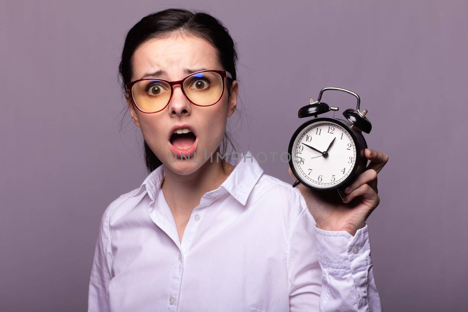 woman manager in a white shirt and glasses holds an alarm clock in her hand, gray studio background by shilovskaya