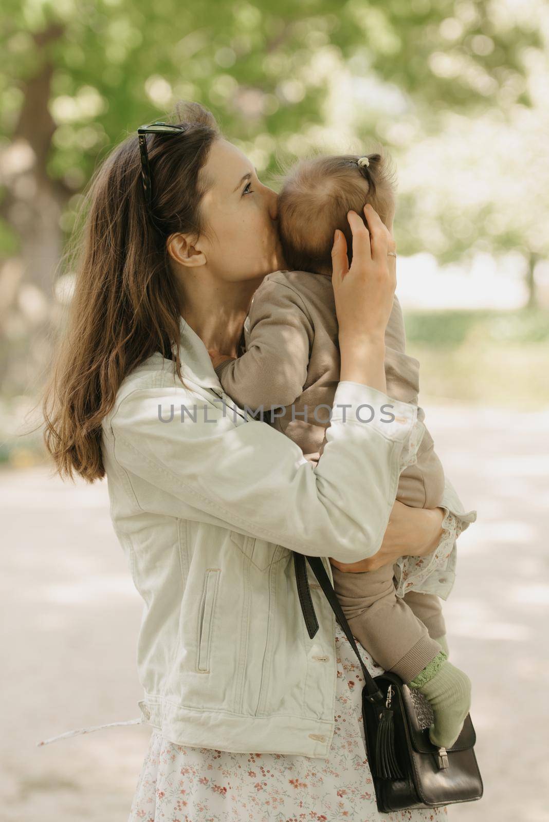 A young mother is holding in his arms her 7-month daughter in the park. A mom is kissing her infant daughter in the woodland.