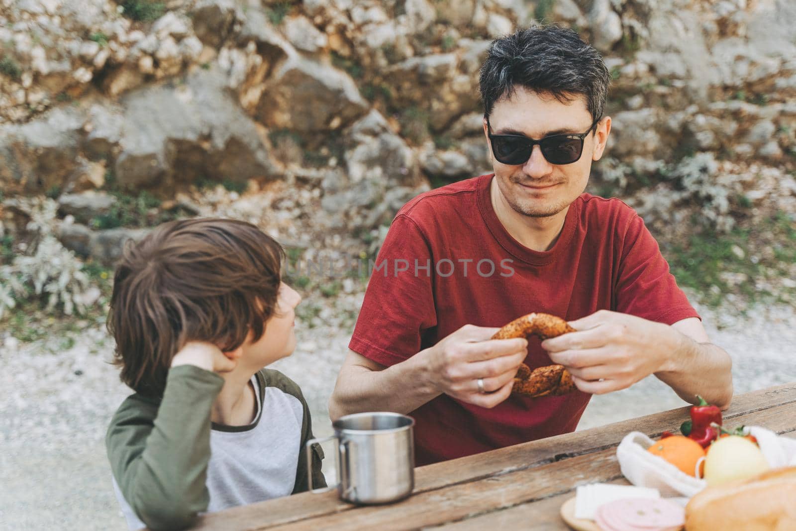 Close-up view of father hugs his school boy son on a family picnic. Child kid and his dad taking a rest and enjoying a picnic while hiking. Boy smiles and bites the bread donut on a picnic by Ostanina