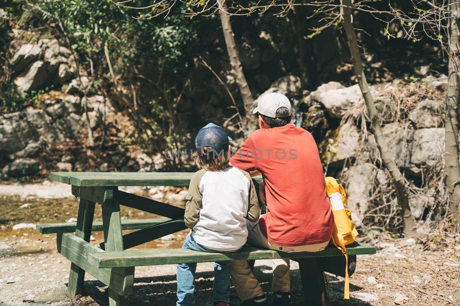 Rear view of tourists school boy and his dad sitting at the wooden table when taking a hike. Child boy and father wearing casual clothes and yellow backpack taking rest on a mountain hike.