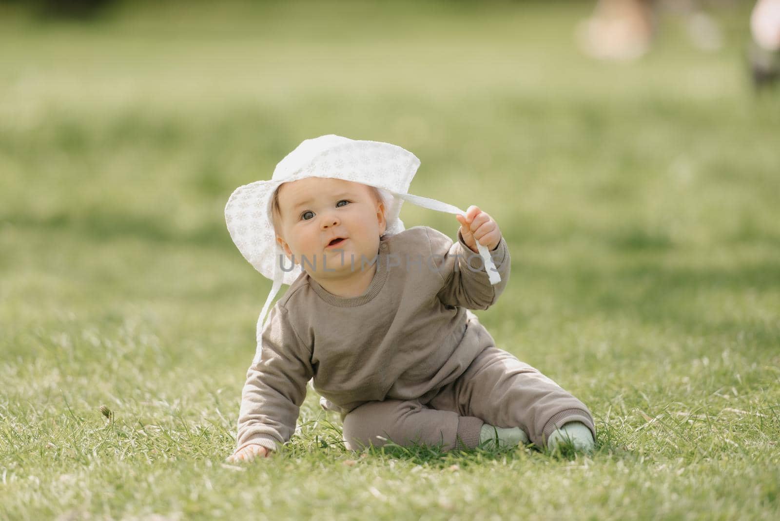 A 7-month child in the white panama hat is having fun in the meadow. An infant girl is crawling on the grass.