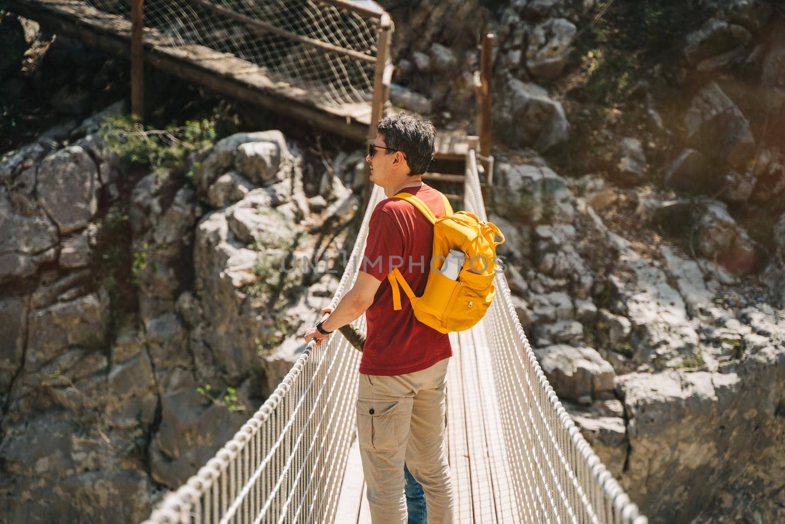 Father and son looking afar from the rope bridge in the mountains when hiking. Casually dressed tourists child school boy and his dad with yellow backpack crossing the canyon through rope bridge.