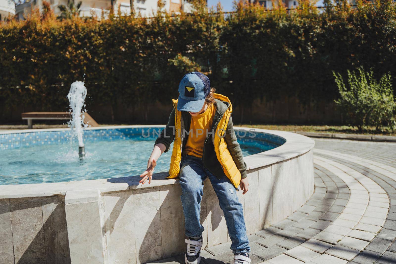 Schoolboy sitting on a fountain in public park during spring sunny day playing with water. Kid in yellow vest chilling in park with water sprinkling in the background. Summer holidays outside.