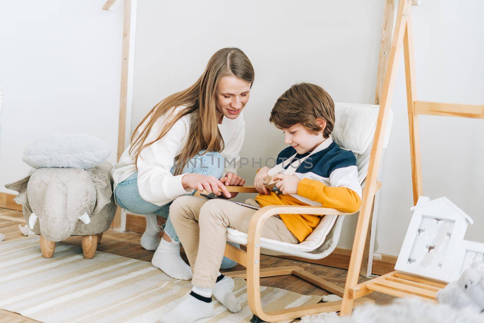 Happy caucasian mother sitting with son in children's room, looking at tablet computer device. Smiling family plays video games with kid boy together. Mom and chilld wathcing on touch screen.
