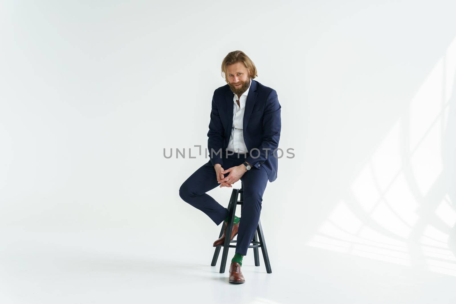 a handsome brutal bearded and long-haired man, a stylish businessman sitting on a chair with a white background. High quality photo