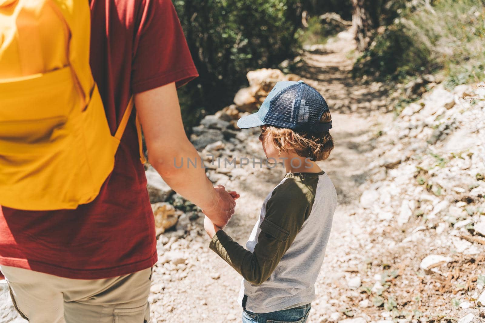 Close-up rear view of tourists school boy and his dad walking a stone footpath in spring forest. Child boy and father wearing casual clothes and yellow backpack while hiking in summer greenwood forest