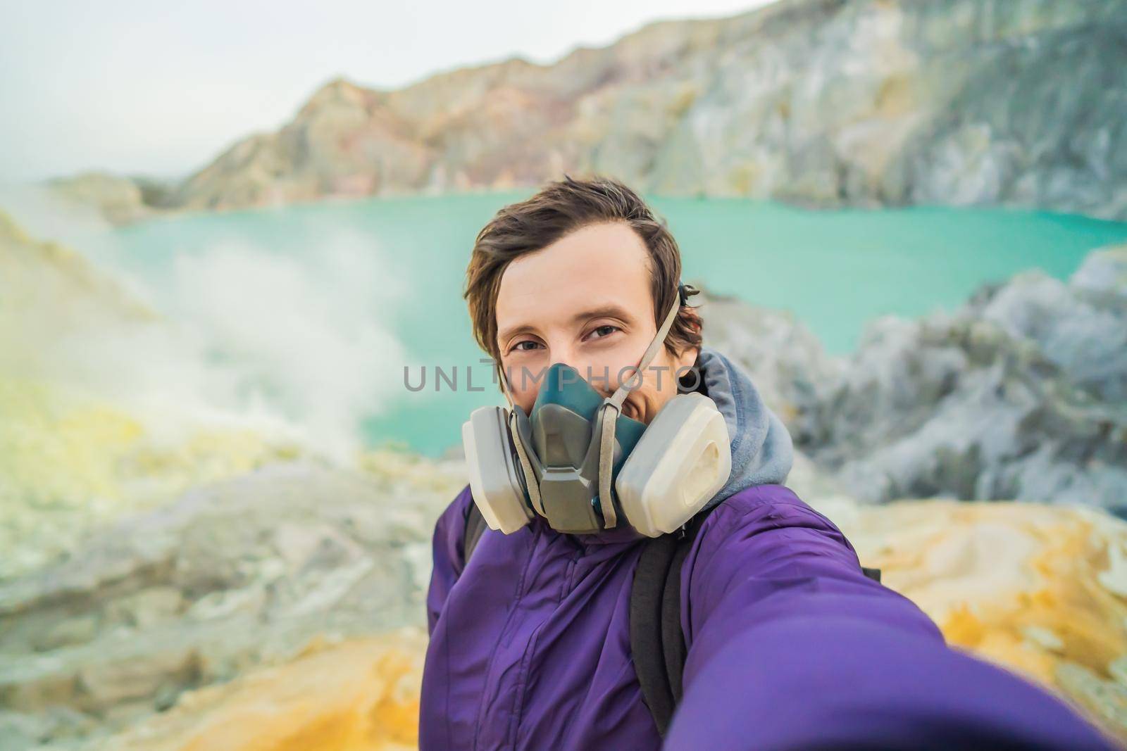 Young man tourist makes a selfie standing at the edge of the crater of the Ijen volcano or Kawah Ijen on the Indonesian language. Famous volcano containing the biggest in the world acid lake and sulfur mining spot at the place where volcanic gasses come from the volcano. He wears a respirator to protect from dangerous volcanic gasses.
