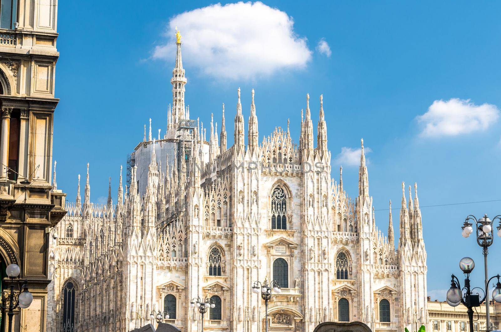 Duomo di Milano cathedral facade with white walls, spires, mouldings and stucco work on Piazza del Duomo square, view from Piazza Mercanti with blue sky background, Milan historical city centre, Italy