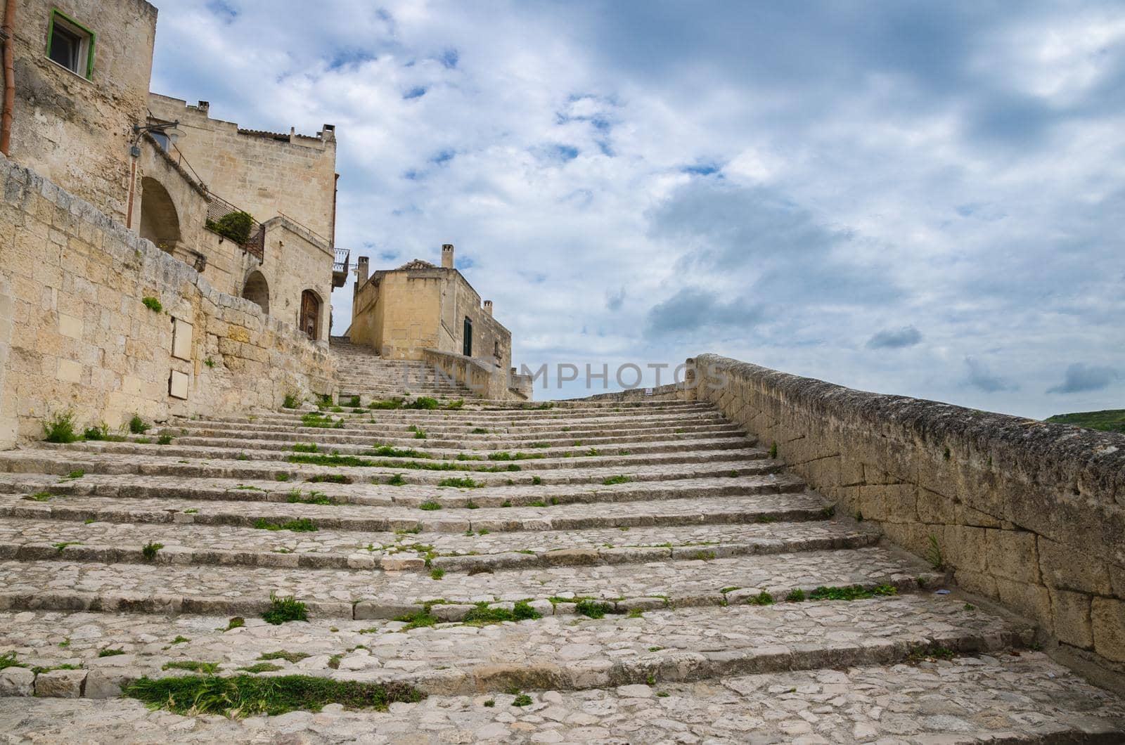 Stone stairs with grass between leading up near old buildings with cloudy sky background in historical centre Sasso Caveoso of ancient town city Sassi di Matera, UNESCO, Basilicata, Southern Italy