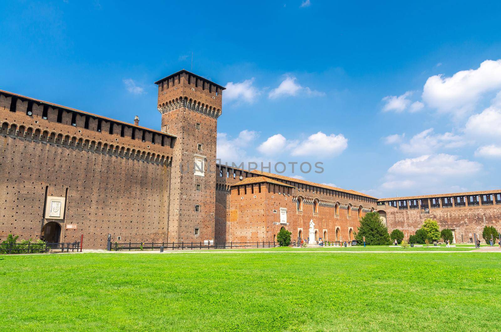 Tower of Porta Giovia and brick walls of old medieval Sforza Castle Castello Sforzesco with green lawn of courtyard and blue sky white clouds background, Milan, Lombardy, Italy