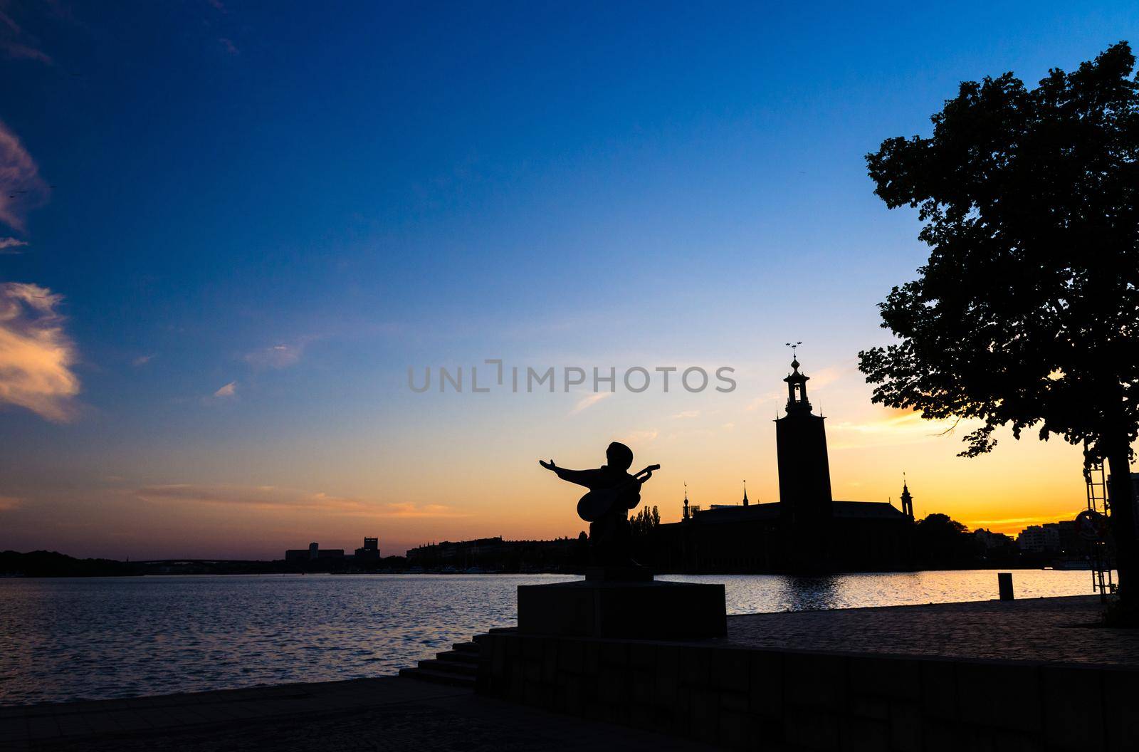 Silhouette of Evert Taube statue monument and Stockholm City Hall (Stadshuset) building Municipal Council on Kungsholmen Island at sunset, dusk, twilight with blue yellow orange sky background, Sweden