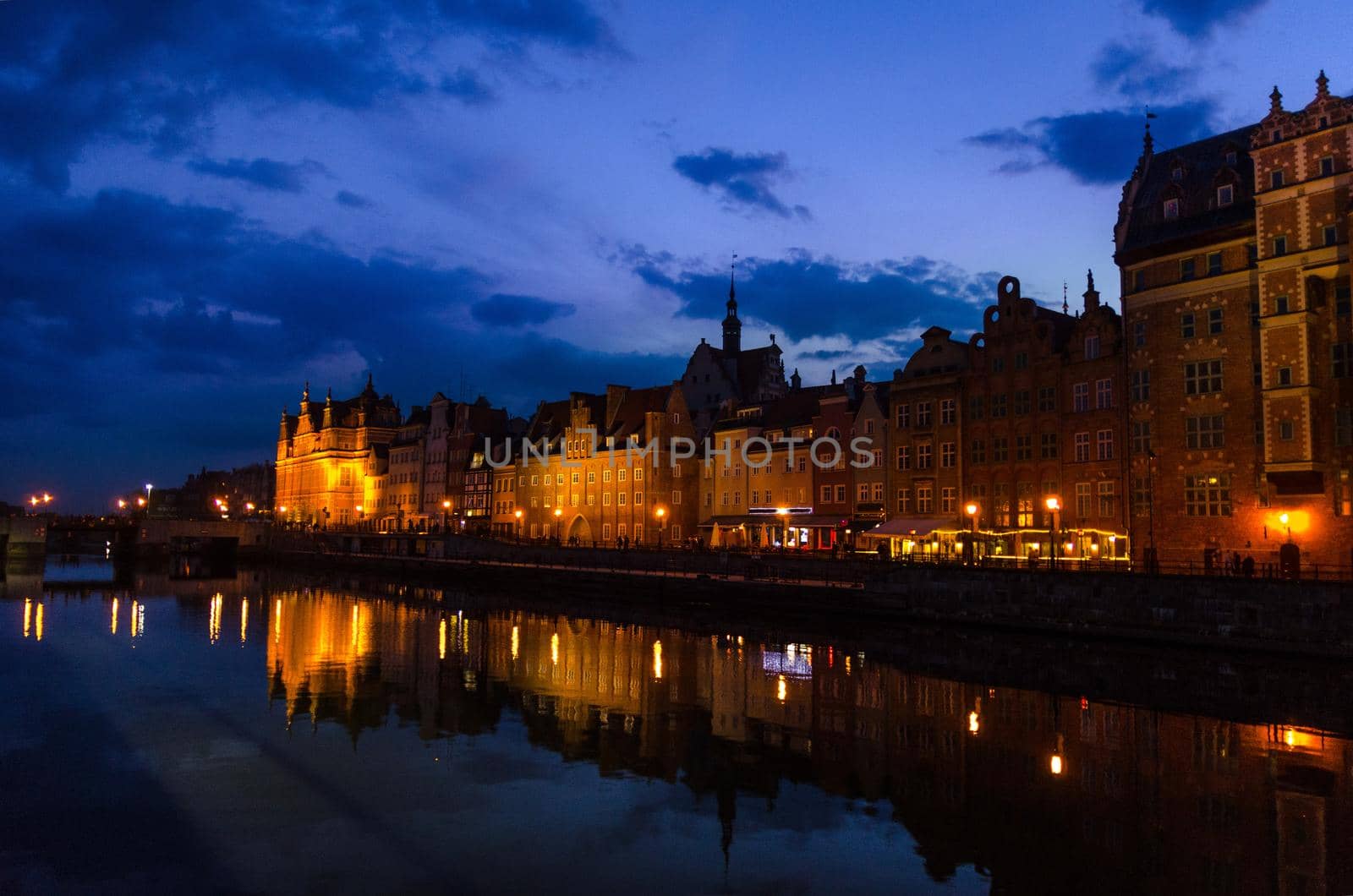 Typical buildings and Green gate Zielona Brama at Long Bridge embankment promenade reflection in Motlawa river water in old historical town centre, street lights dark blue sky evening, Gdansk, Poland