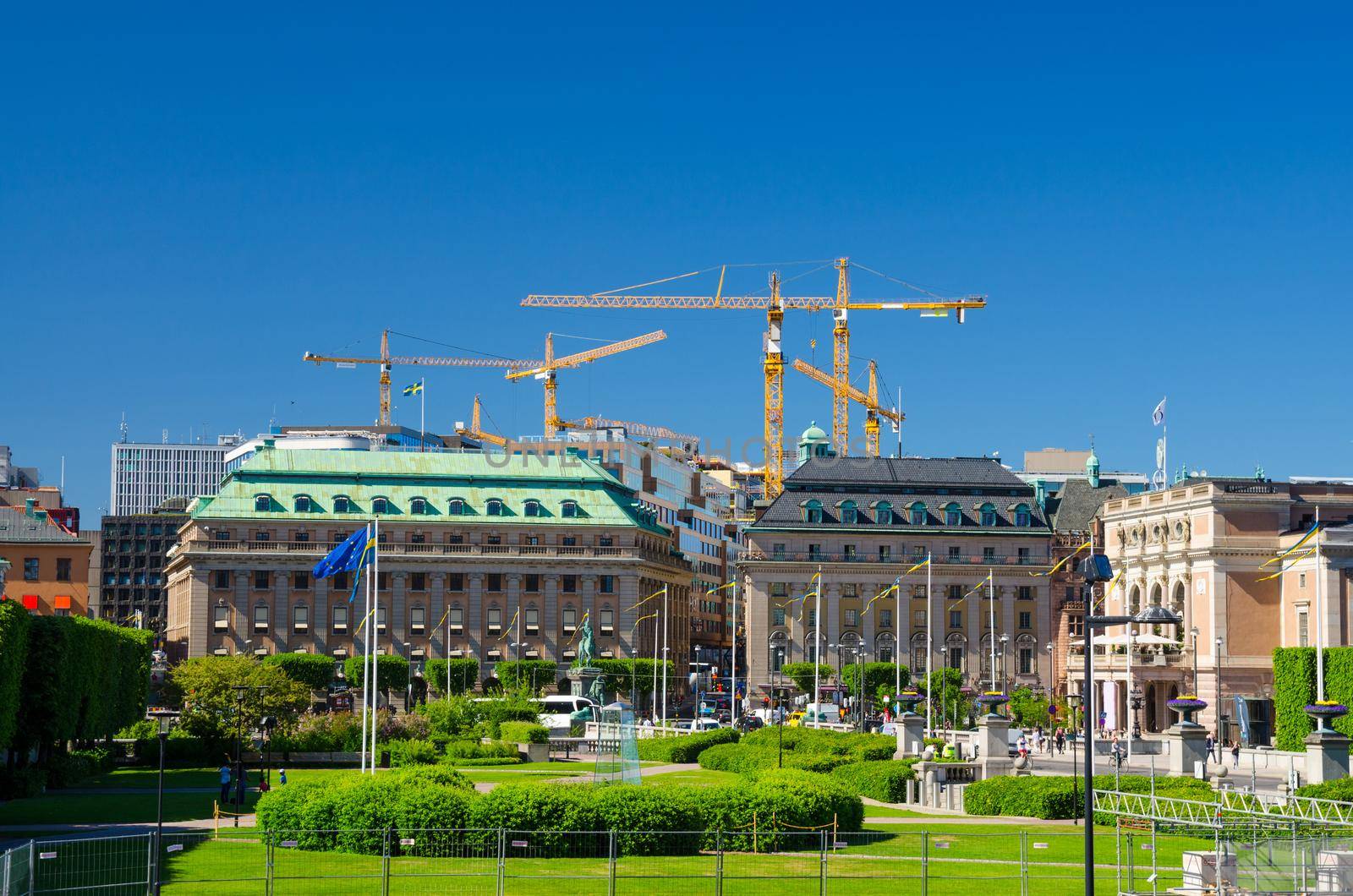 Riksplan green grass lawn, bushes and street with national flags on Sodermalm island, Gustav Adolfs torg, Royal Swedish Opera house building, blue sky with yellow cranes background, Stockholm, Sweden