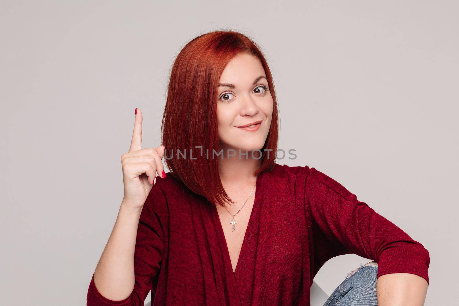 Portrait of young beautiful woman on gray background smiling at camera. Pretty red haired girl in bordo blouse and jeans thinking about something. Concept of new idea and thinking.