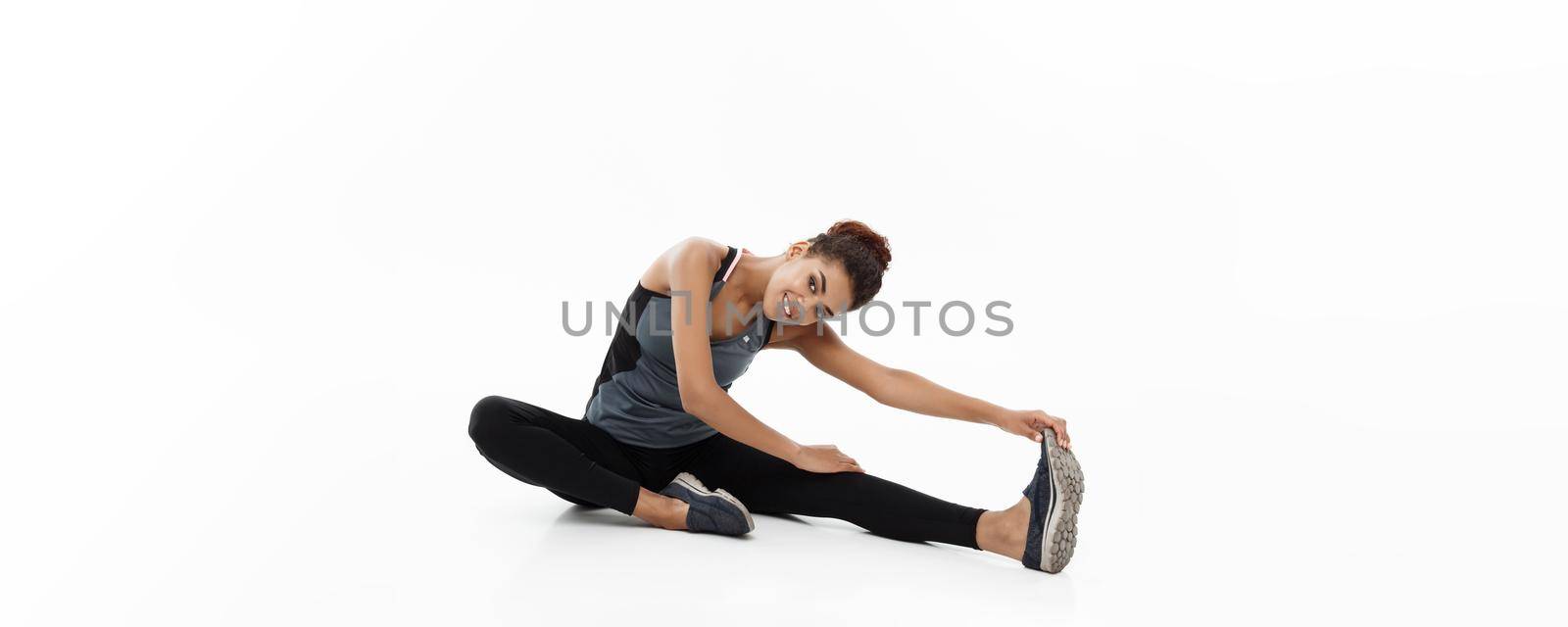 Sport, training, lifestyle and Fitness concept - portrait of beautiful happy African American woman stretching leg while sitting. Isolated on white studio background. by Benzoix