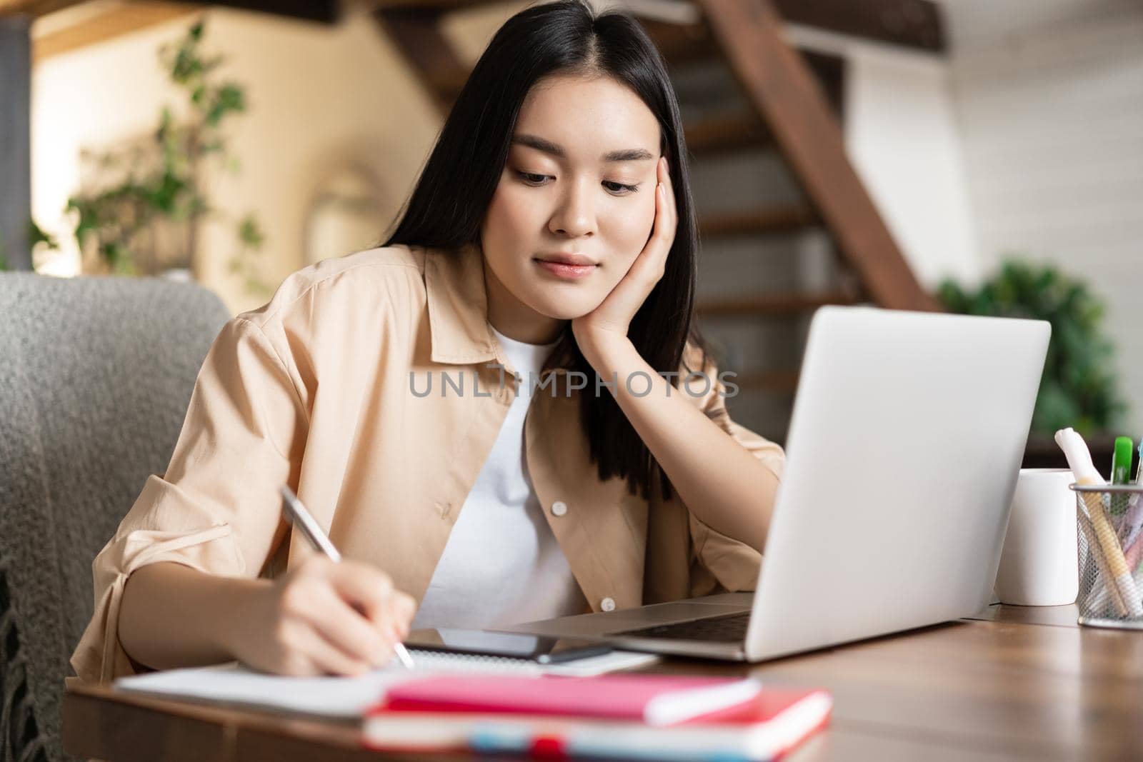 Young asian girl taking notes, writing and doing homework, sitting near laptop. Korean woman working from home with computer pc.