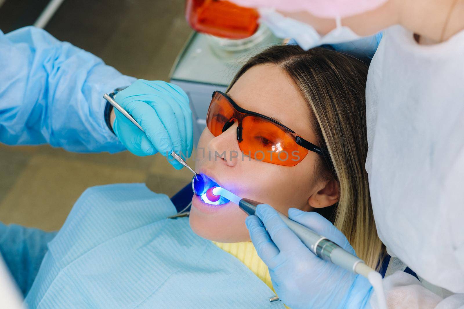 A young beautiful girl in dental glasses treats her teeth at the dentist with ultraviolet light. filling of teeth by Lobachad