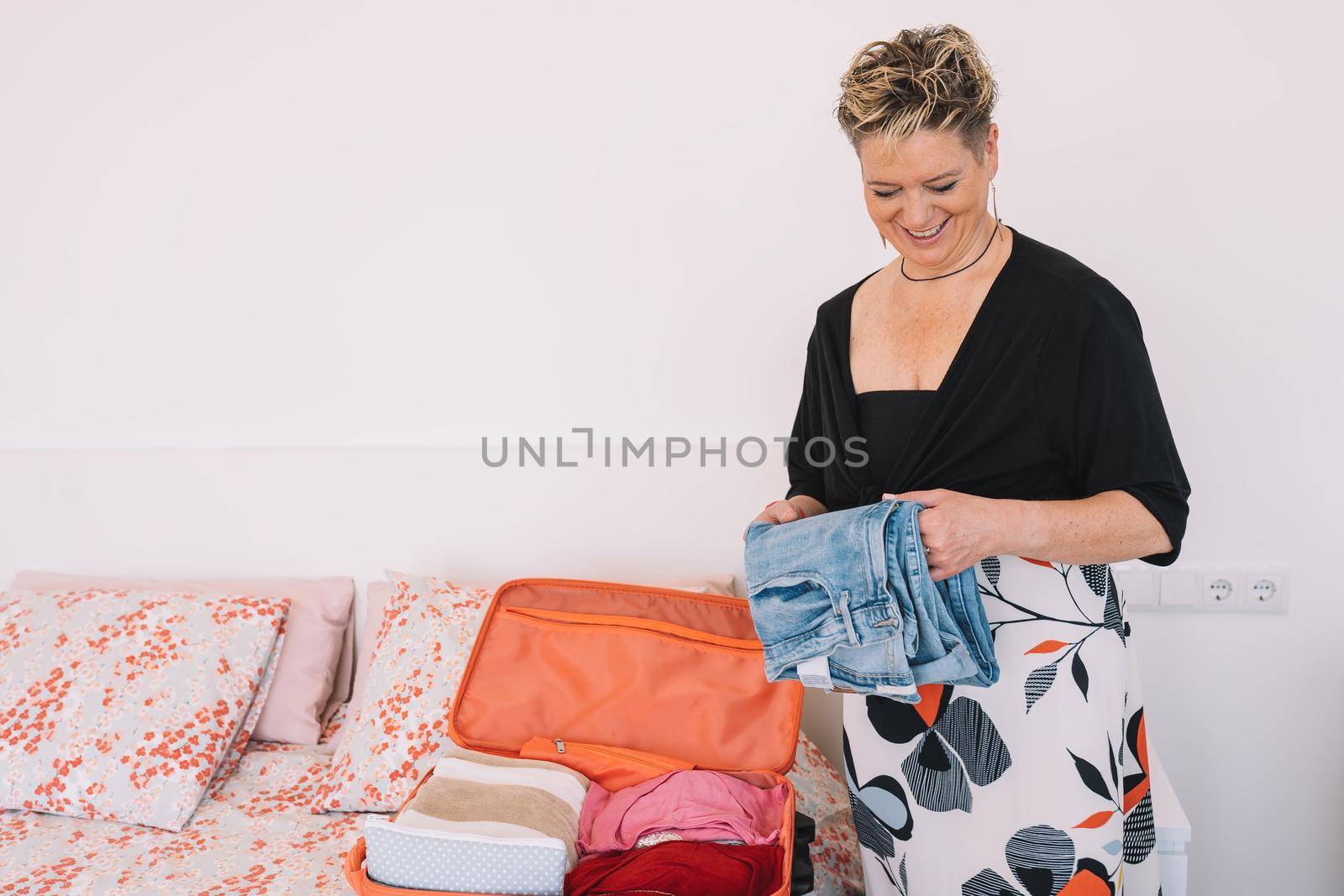 mature woman folding clothes to prepare her luggage. woman owner of establishment preparing her suitcase for business trip. copy space. concept of holidays, travel. bedroom with natural light, uncluttered environment. with white walls.