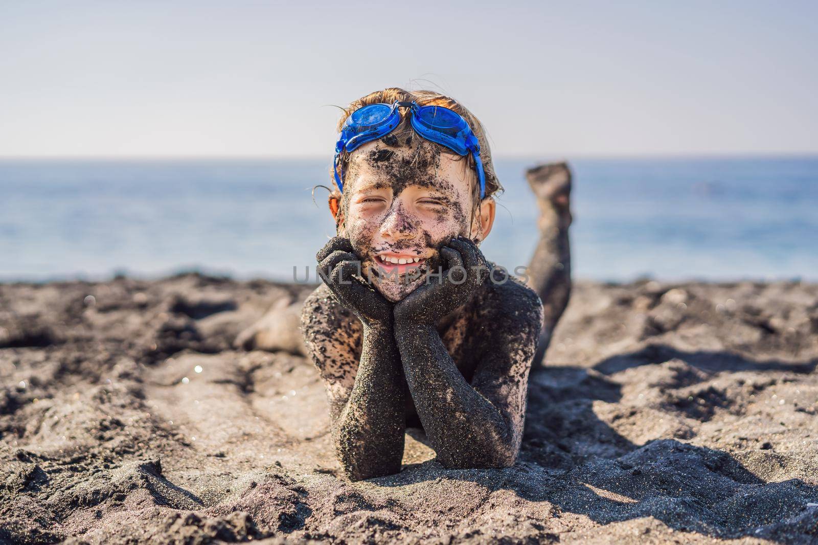 Black Friday concept. Smiling boy with dirty Black face sitting and playing on black sand sea beach before swimming in ocean. Family active lifestyle, and water leisure on summer vacation with kids. Black Friday, sales of tours and airline tickets or goods.