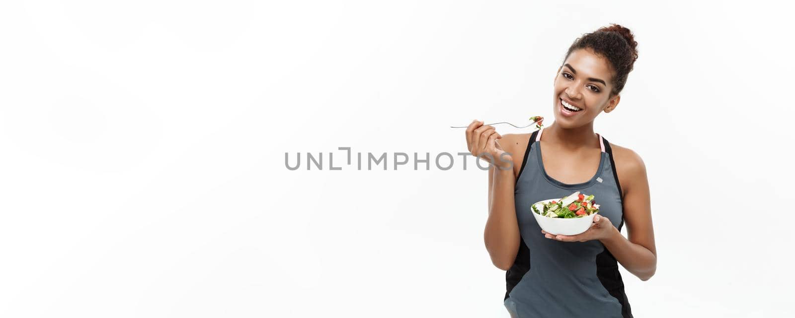 Healthy and Fitness concept - Beautiful American African lady in fitness clothes on diet eating fresh salad. Isolated on white background. by Benzoix