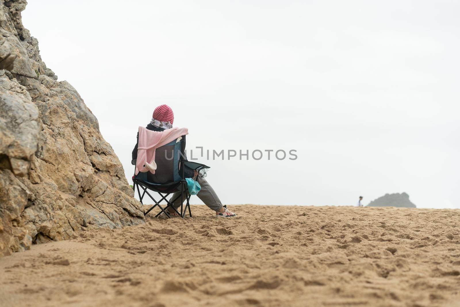 Thoughtful elderly woman sitting touristic chair on sea beach looking horizon Loneliness and old age concept Autumn of life Cold weather warm clothes