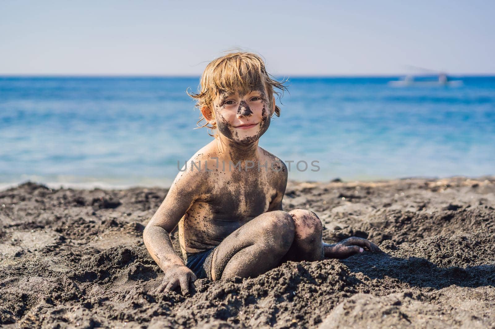 Black Friday concept. Smiling boy with dirty Black face sitting and playing on black sand sea beach before swimming in ocean. Family active lifestyle, and water leisure on summer vacation with kids. Black Friday, sales of tours and airline tickets or goods by galitskaya