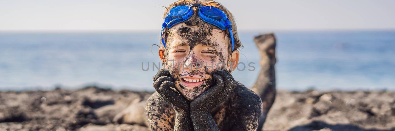 BANNER, LONG FORMAT Black Friday concept. Smiling boy with dirty Black face sitting and playing on black sand sea beach before swimming in ocean. Family active lifestyle, and water leisure on summer vacation with kids. Black Friday, sales of tours and airline tickets or goods.
