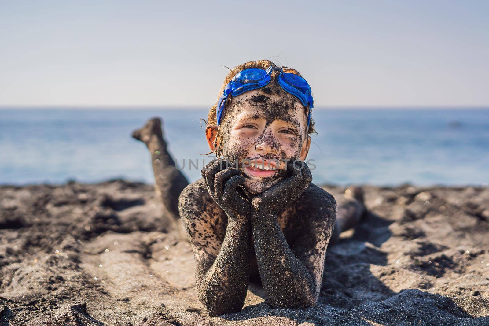 Black Friday concept. Smiling boy with dirty Black face sitting and playing on black sand sea beach before swimming in ocean. Family active lifestyle, and water leisure on summer vacation with kids. Black Friday, sales of tours and airline tickets or goods.